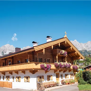 A traditional, wood-clad house with blooming flowers on the balcony. Majestic mountains and a clear sky can be seen in the background.