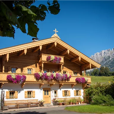A traditional wooden house with colorful flower pots and a clear blue sky. Impressive mountains are visible in the background.