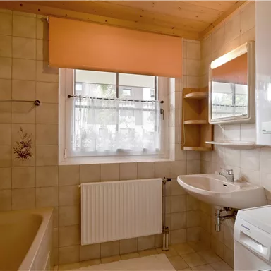 Eine helle Badezimmeransicht mit Badewanne, Waschbecken und Waschmaschine. Die Wände sind gefliest und es gibt ein Fenster mit einem Gardinenaufsatz.