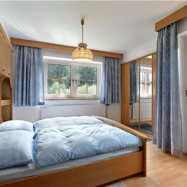 Ein gemütliches Schlafzimmer mit einem großen Bett und blauen Bettdecken. Der Raum hat große Fenster und Holzregal mit einem kleinen Tisch.