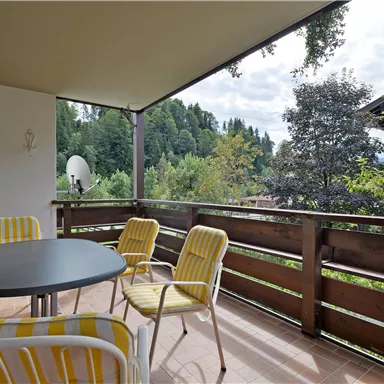 Ein gemütlicher Balkon mit einem Tisch und gelben Stühlen. Im Hintergrund sieht man eine grüne Landschaft und Bäume.