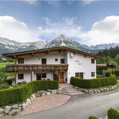 A charming house in the mountains with a well-kept garden. In the background, majestic mountains and a clear sky can be seen.