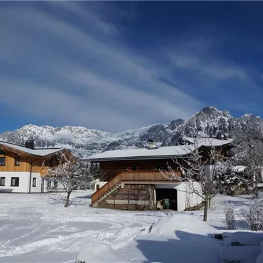 A winter landscape with snow-covered houses and mountains in the background. The sky is blue with some clouds.