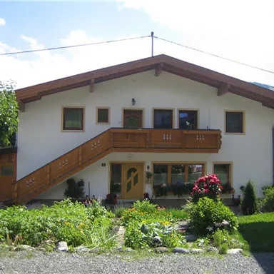 Ein hübsches Haus mit einem Holzbalkon und Pflanzen im Vordergrund. Die Umgebung ist grün und bergig.