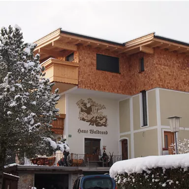 Ein modernes Haus im alpinen Stil, umgeben von schneebedeckten Bäumen. Die Fassade ist teilweise mit Holz verkleidet und hat einen dekorativen Schriftzug.