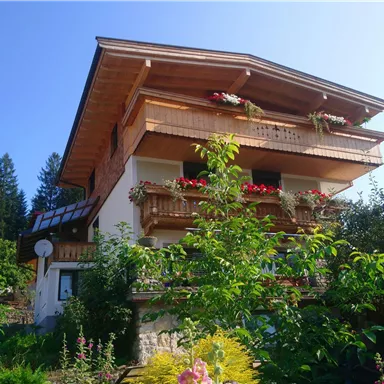 Ein charmantes Haus mit mehreren Balkonen, umgeben von blühenden Pflanzen und Bäumen. Der Himmel ist klar und sonnig, was die idyllische Atmosphäre verstärkt.
