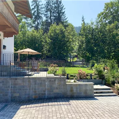 Eine gepflegte Terrasse mit einem Tisch und einem Sonnenschirm. Im Hintergrund sind grüne Bäume und ein schöner Garten.