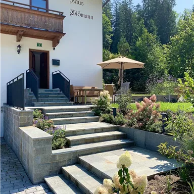 Ein einladender Eingangsbereich mit Steinstufen und grünem Garten. Im Hintergrund steht eine helle Fassade mit einem Balkon.