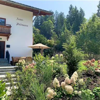 Ein charmantes Haus mit einem Balkon und einem schönen Garten voller Blumen und Pflanzen. Im Hintergrund sind Bäume und eine sonnige Atmosphäre zu sehen.
