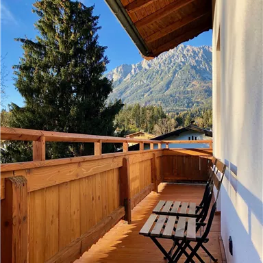 Ein gemütlicher Balkon mit Holzgeländer und zwei Stühlen. Im Hintergrund sind majestätische Berge und ein klarer blauer Himmel sichtbar.