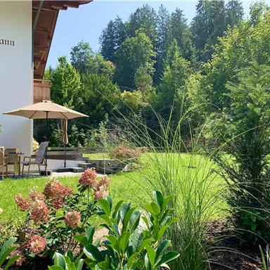 Ein schöner Garten mit bunten Blumen und grünen Pflanzen. Im Hintergrund steht ein Haus mit einer Terrasse und einem Sonnenschirm.