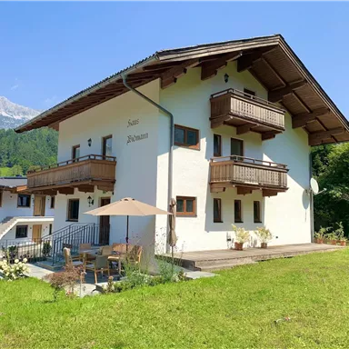 Ein schönes Haus mit weißen Wänden und Balkon im alpinen Stil. Es liegt in einer grünen Umgebung mit Bergen im Hintergrund und einem Garten.