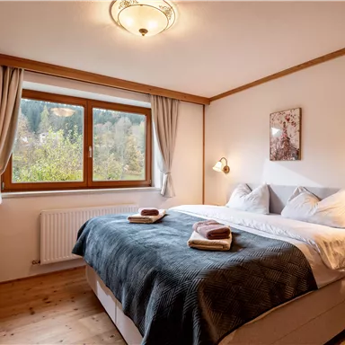 Ein gemütliches Schlafzimmer mit einem großen Boxspringbett und einem Fenster, das auf die Natur blickt. Es ist hell und einladend, mit warmer Holzdecke und sanften Farben.