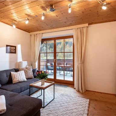 Ein gemütliches Wohnzimmer mit einer grauen Couch und einem Tisch. Große Fenster mit Vorhängen bieten einen Blick auf die Terrasse .