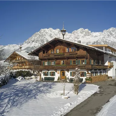 Ein traditionelles, holzverziertes Bauernhaus in einer schneebedeckten Landschaft. Im Hintergrund erheben sich majestätische Berge unter klarem blauen Himmel.