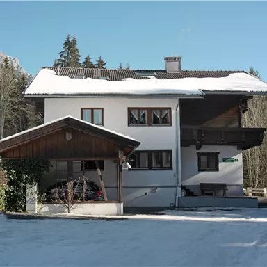 Ein zweigeschossiges Wohnhaus mit einem schneebedeckten Dach. Im Hintergrund sind verschneite Berge und ein klarer blauer Himmel zu sehen.