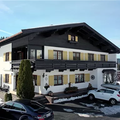 A traditional alpine house with black wood and yellow shutters. It is located in a snowy environment with trees in the background.