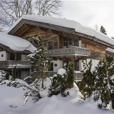 Ein rustikales Holzhaus im Schnee, umgeben von hohen Nadelbäumen. Der Himmel ist klar und es ist eine friedliche Winterlandschaft zu sehen.