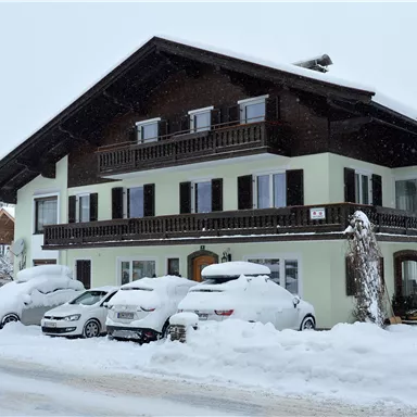 Ein großes, zweigeschossiges Haus im winterlichen Schnee. Die Autos vor dem Haus sind unter einer dicken Schneeschicht verborgen.