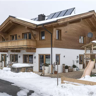 Ein modernes Holzhaus mit einem Balkon und einer Rutsche im Schnee. Im Hintergrund sind weitere Häuser und schneebedeckte Landschaft zu sehen.