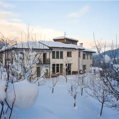 A beautiful house in the snow, surrounded by snow-covered trees. The sky is clear and showcases a wintry landscape.