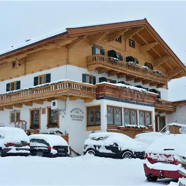 Ein traditionelles Holzhaus im Schnee. Parkierte Autos sind unter einer dicken Schneedecke verborgen.