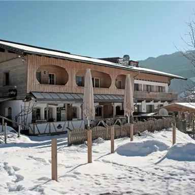 A charming wooden house in the snow with large windows and sunshades. The surroundings are surrounded by mountains and trees.