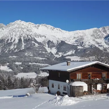 Ein rustikales Holzhaus im Schnee, umgeben von majestätischen Bergen. Der Himmel ist klar und blau.