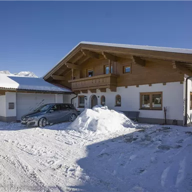 A charming wooden house in the snow with a red vehicle in front. The surroundings are mountainous and the sun is shining.