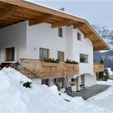 A modern house with a wooden porch in a snow-covered landscape. Impressive mountains can be seen in the background.