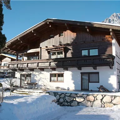 Ein gemütliches Chalet im Schnee mit Holzverkleidung und Balkonen. Im Hintergrund sind schneebedeckte Berge zu sehen.