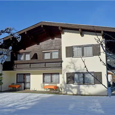 A beautiful house in the snow with a clear blue sky.  
In front of the house are orange chairs on the terrace.