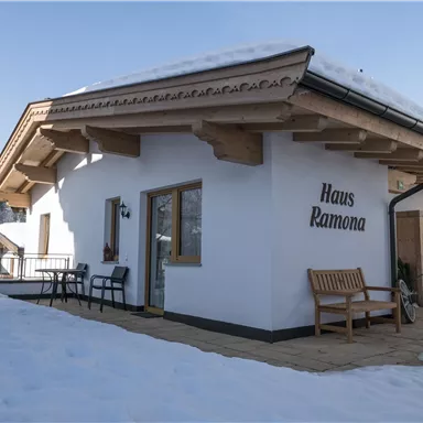 A cozy, white holiday home named "Haus Ramona". It is located in a snowy environment and has a terrace with seating furniture.