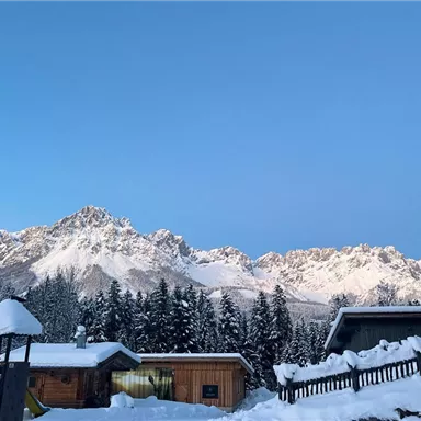 Eine verschneite Landschaft mit Bergen im Hintergrund und schneebedeckten Hütten im Vordergrund. Der Himmel ist klar und blau.