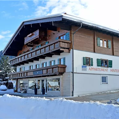 A modern apartment building in alpine style, surrounded by snow. The facade is bright and inviting with several windows and balconies.