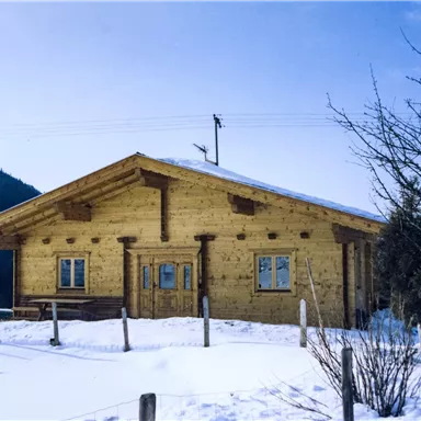 Ein gemütliches Holzhaus in einer verschneiten Landschaft. Im Hintergrund sind Berge und blauer Himmel zu sehen.
