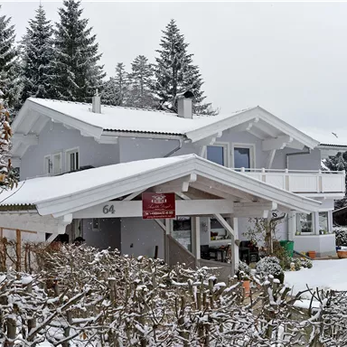 A charming house in the snow, surrounded by snow-covered trees. The sky is gray and overcast, creating a tranquil winter atmosphere.