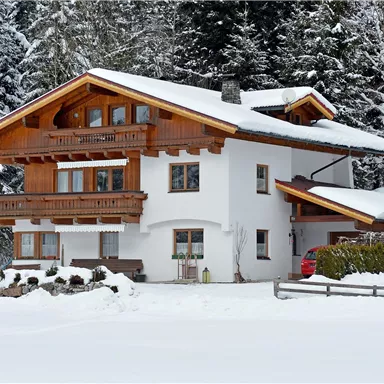 A beautiful, wintery house with wooden cladding, surrounded by snow-covered trees. The snow-covered garden and quiet ambiance convey a cozy atmosphere.
