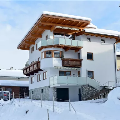 Ein modernes Haus im Schnee, umgeben von anderen Gebäuden. Der Balkon und die Holzverkleidung verleihen dem Gebäude einen charmanten alpinen Stil.