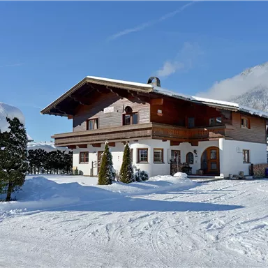 Ein gemütliches Holzhaus im Schnee mit einem klaren blauen Himmel. Umgeben von einer winterlichen Landschaft mit schneebedeckten Bäumen.