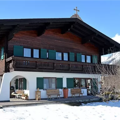 Ein traditionelles Holzhaus mit grünen Fensterläden und einem Kreuz auf dem Dach. Es steht in einer schneebedeckten Landschaft mit Bergen im Hintergrund.