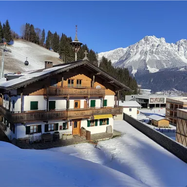 A traditional wooden house in a snowy landscape. In the background, there are high mountains and a cable car.