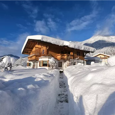 Ein gemütliches, zweistöckiges Holzhaus in verschneiter Landschaft. Hoher Schnee und blauer Himmel umgeben die idyllische Szene.