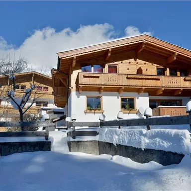A picturesque winter landscape with snow-covered cottages. The sky is blue and the mountains are visible in the background.