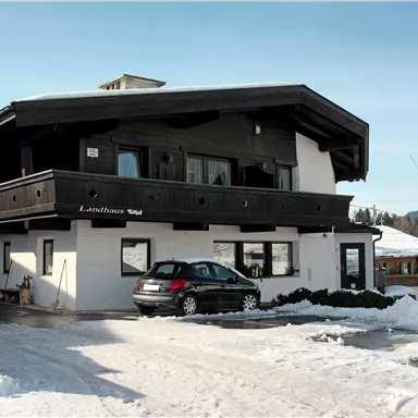 Ein gemütliches, mehrstöckiges Haus im Schnee mit einem Auto davor. Im Hintergrund sind schneebedeckte Berge zu sehen.