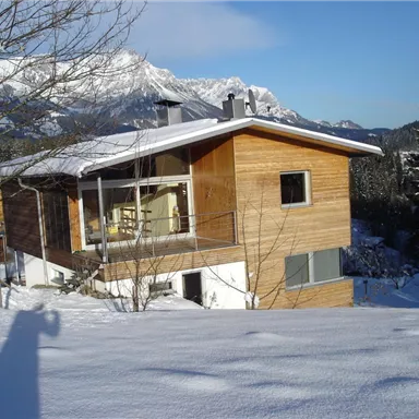 Ein modernes Holzhaus im Schnee, umgeben von Bergen. Der klare Himmel und die schneebedeckte Landschaft schaffen eine ruhige Winteratmosphäre.