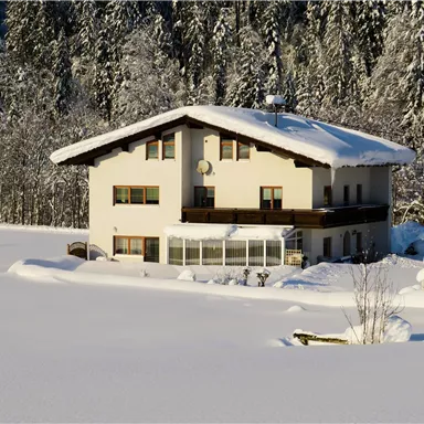 Ein gemütliches Haus im Schnee, umgeben von einem weißen Winterwunderland. Die Bäume im Hintergrund sind mit einer dicken Schicht Schnee bedeckt.