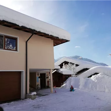 A modern house in the snow with a large snow pile on the roof. In the background, there are snow-covered mountains and a clear blue sky.