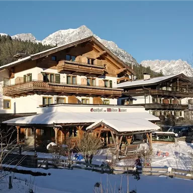 A traditional Austrian hotel in winter with snow-covered grounds. In the background, there are mountains and clear blue skies.