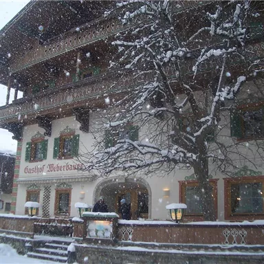 Ein traditionelles Gasthaus im Schnee, umgeben von winterlichen Bäumen. Große Schneeflocken fallen auf die charmante Fassade.